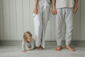 A toddler crawls on the floor while parents stand holding hands in a cozy home setting wearing casual clothes.